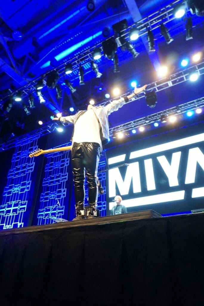 Guitarist MIYAVI onstage at Anime Boston 2019, standing with his arms outstretched and his head flung back. He's wearing a loose, white shirt and black pants with a white stripe along the side.