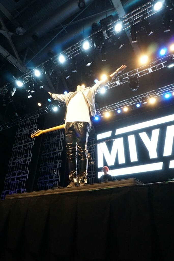 Guitarist MIYAVI onstage at Anime Boston 2019, standing with his arms outstretched and his head flung back. He's wearing a loose, white shirt and black pants with a white stripe along the side.