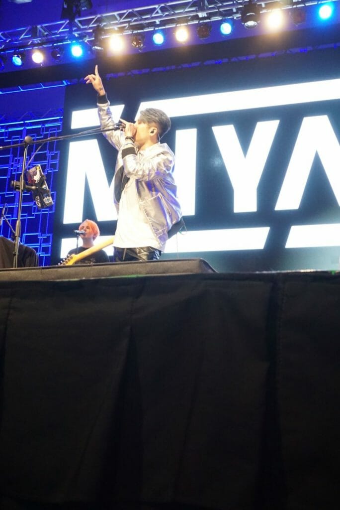 MIYAVI onstage at Anime Boston 2019, with his hand raised as he sings into a mic. He's wearing a loose, white shirt and black pants with a white stripe along the side.