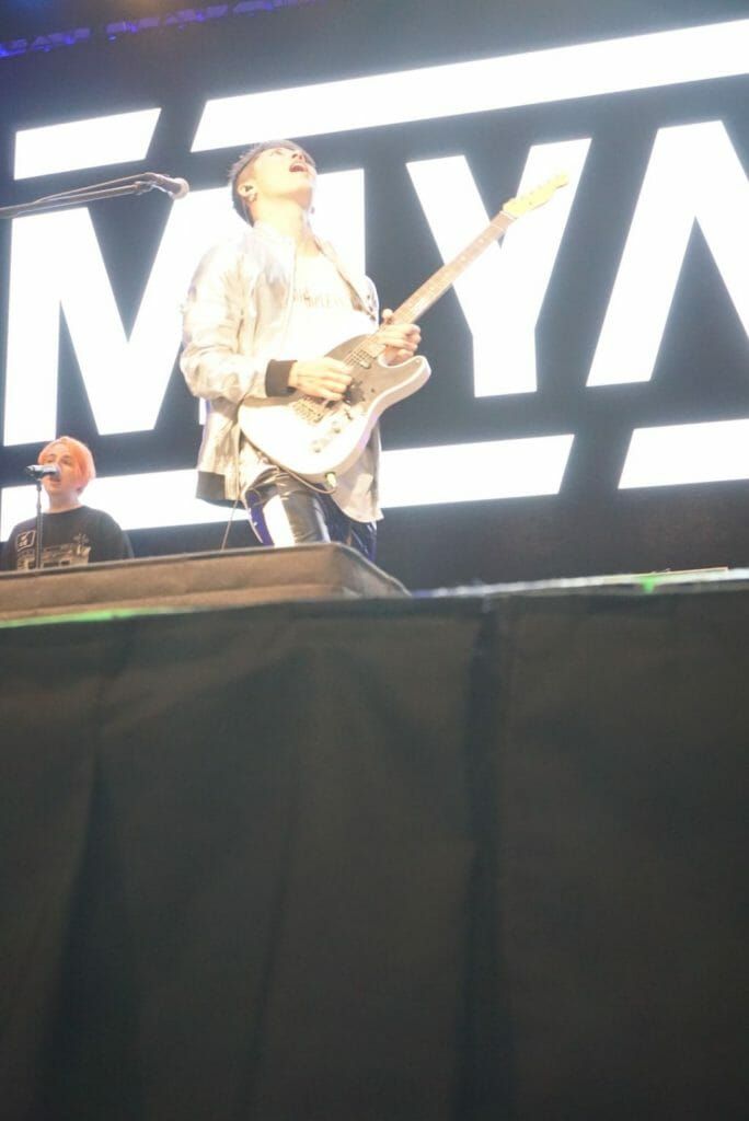 Guitarist MIYAVI onstage at Anime Boston 2019, triumphantly crying out as he plays his his guitar. He's wearing a loose, white shirt and black pants with a white stripe along the side.