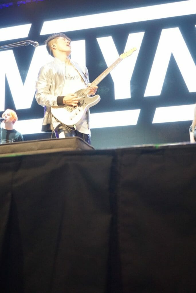 Guitarist MIYAVI onstage at Anime Boston 2019, triumphantly crying out as he plays his his guitar. He's wearing a loose, white shirt and black pants with a white stripe along the side.