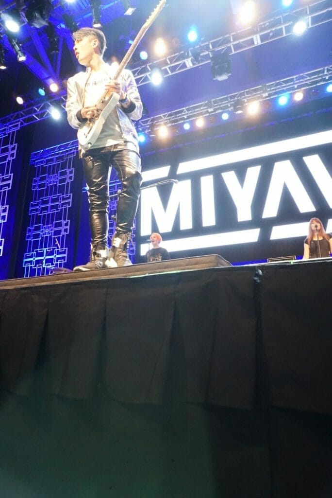 Guitarist MIYAVI onstage at Anime Boston 2019, looking out to the audience as he plays his guitar. He's wearing a loose, white shirt and black pants with a white stripe along the side.