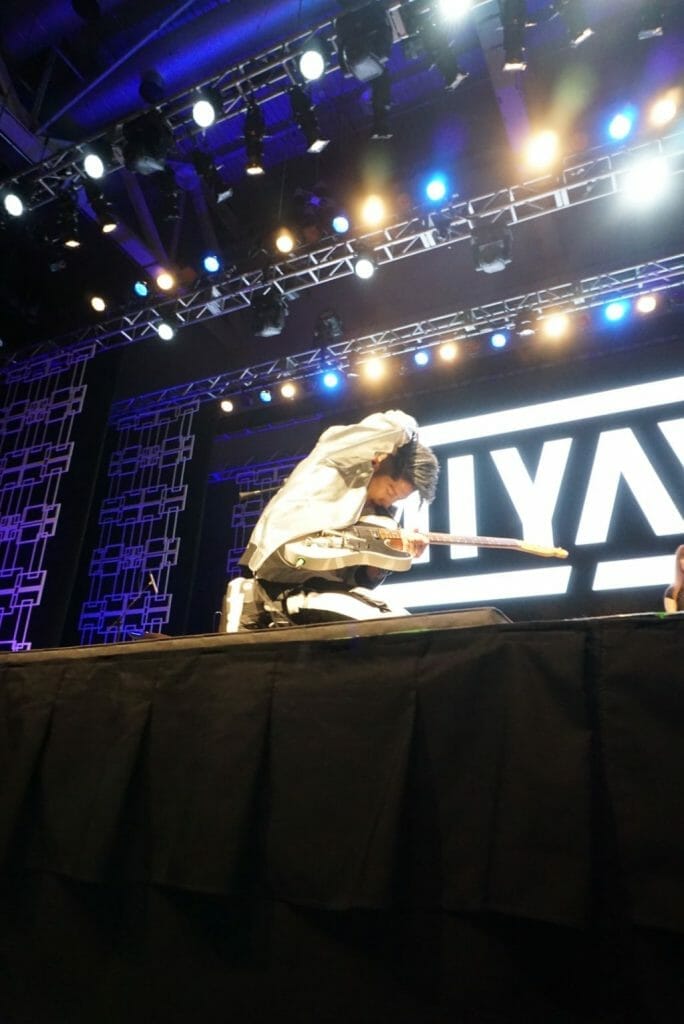 Guitarist MIYAVI onstage at Anime Boston 2019, crouching onstage. He's wearing a loose, white shirt and black pants with a white stripe along the side.