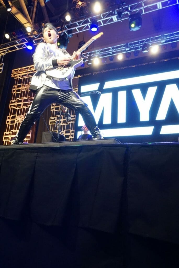 Guitarist MIYAVI onstage at Anime Boston 2019, singing and posing as he plays his guitar. He's wearing a loose, white shirt and black pants with a white stripe along the side.