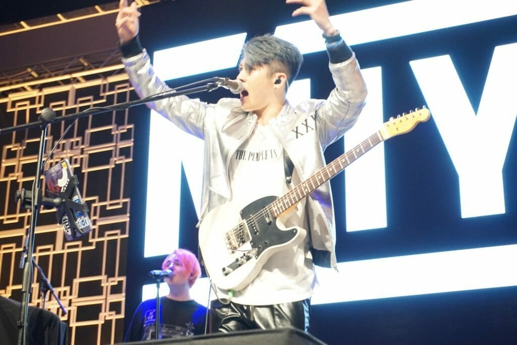 Guitarist MIYAVI onstage at Anime Boston 2019, standing behind the mic as he points out to the audience. He's wearing a loose, white shirt and black pants with a white stripe along the side.