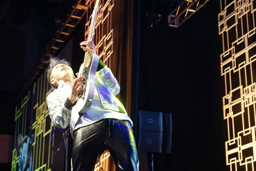 Guitarist MIYAVI onstage at Anime Boston 2019, throwing his head back as he shreds on his guitar. He's wearing a loose, white shirt and black pants with a white stripe along the side.