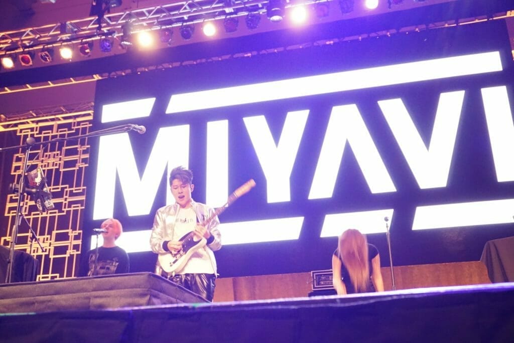 Guitarist MIYAVI onstage at Anime Boston 2019, looking down as he plays his guitar. He's wearing a loose, white shirt and black pants with a white stripe along the side.