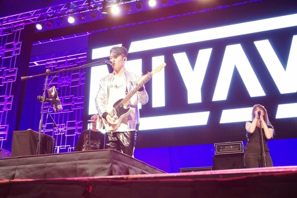MIYAVI onstage at Anime Boston 2019, singing as he plays his guitar. He's wearing a loose, white shirt and black pants with a white stripe along the side.