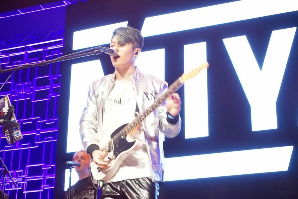 MIYAVI onstage at Anime Boston 2019, singing as he plays his guitar. He's wearing a loose, white shirt and black pants with a white stripe along the side.