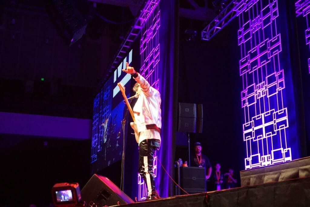 MIYAVI onstage at Anime Boston 2019, pointing out toward stage left. He's wearing a loose, white shirt and black pants with a white stripe along the side.