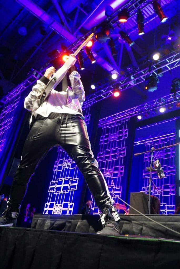 MIYAVI onstage at Anime Boston 2019, leaning back as he shreds on his guitar. He's wearing a loose, white shirt and black pants with a white stripe along the side.