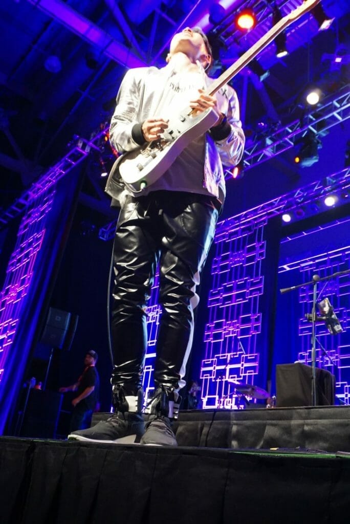 MIYAVI onstage at Anime Boston 2019, standing upright s he looks toward the ceiling. He's wearing a loose, white shirt and black pants with a white stripe along the side.