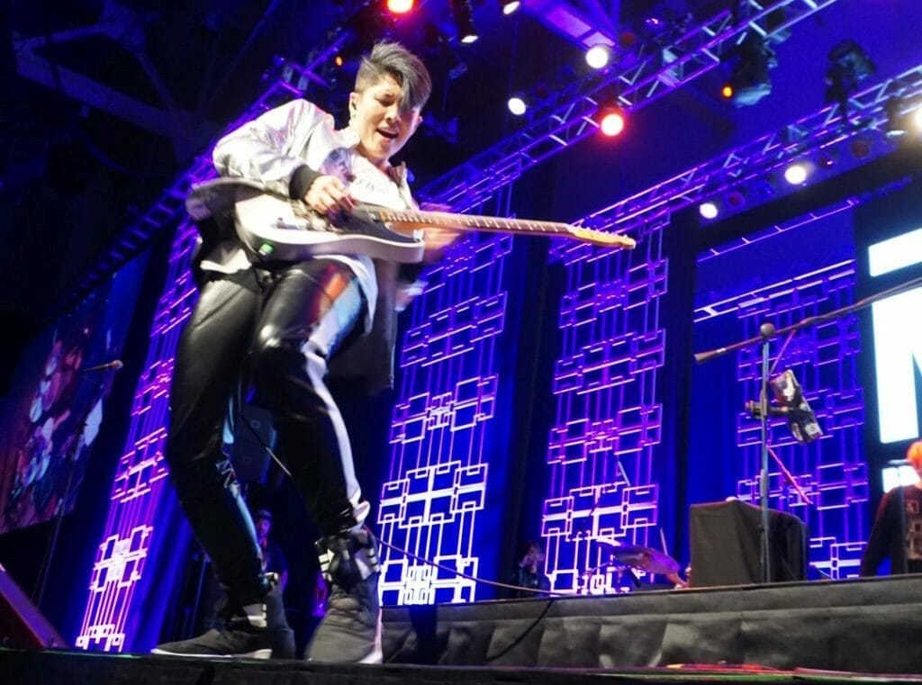 Guitarist MIYAVI onstage at Anime Boston 2019, grinning ear to ear as he plays his guitar. He's wearing a loose, white shirt and black, leather pants.