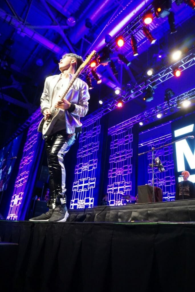 MIYAVI onstage at Anime Boston 2019, turning toward stage right. He's wearing a loose, white shirt and black pants with a white stripe along the side.