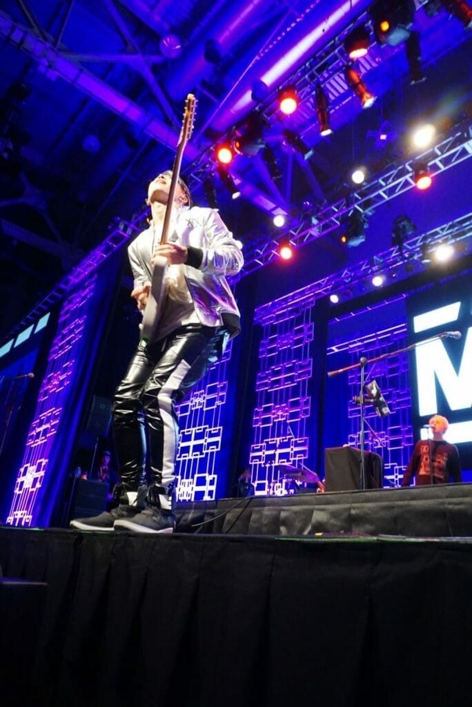 MIYAVI onstage at Anime Boston 2019, grinning ear to ear as he shreds on his guitar. He's wearing a loose, white shirt and black pants with a white stripe along the side.