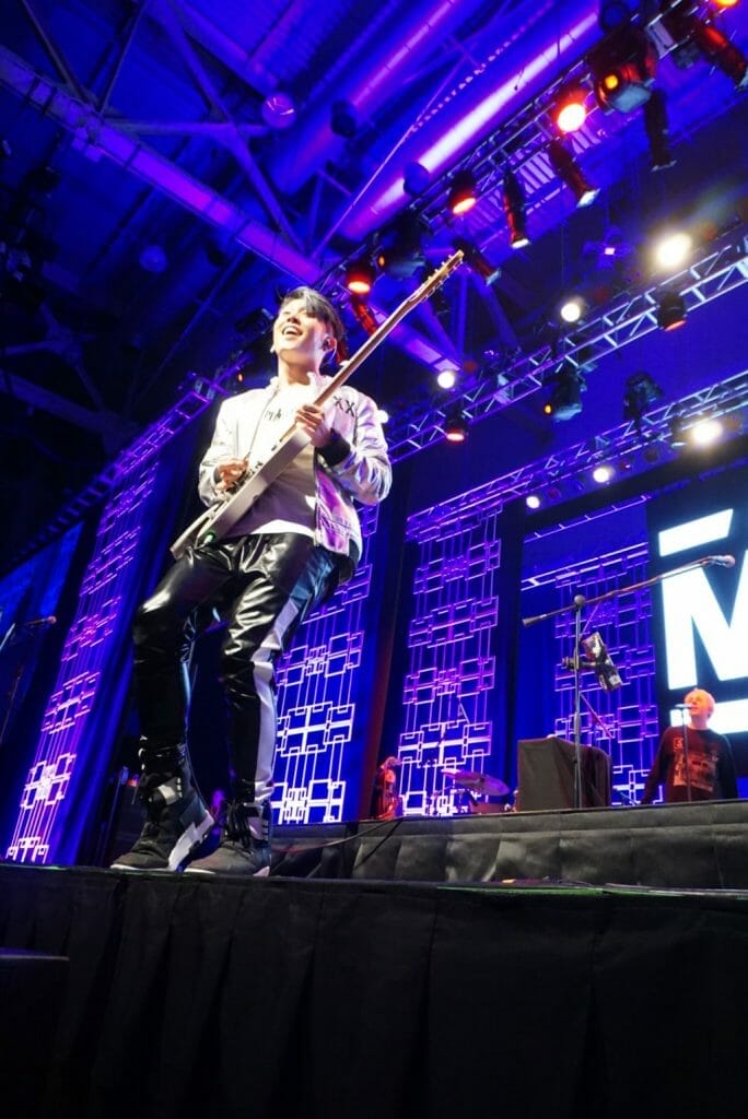 Guitarist MIYAVI onstage at Anime Boston 2019, grinning ear to ear as he shreds on his guitar. He's wearing a loose, white shirt and black pants with a white stripe along the side.