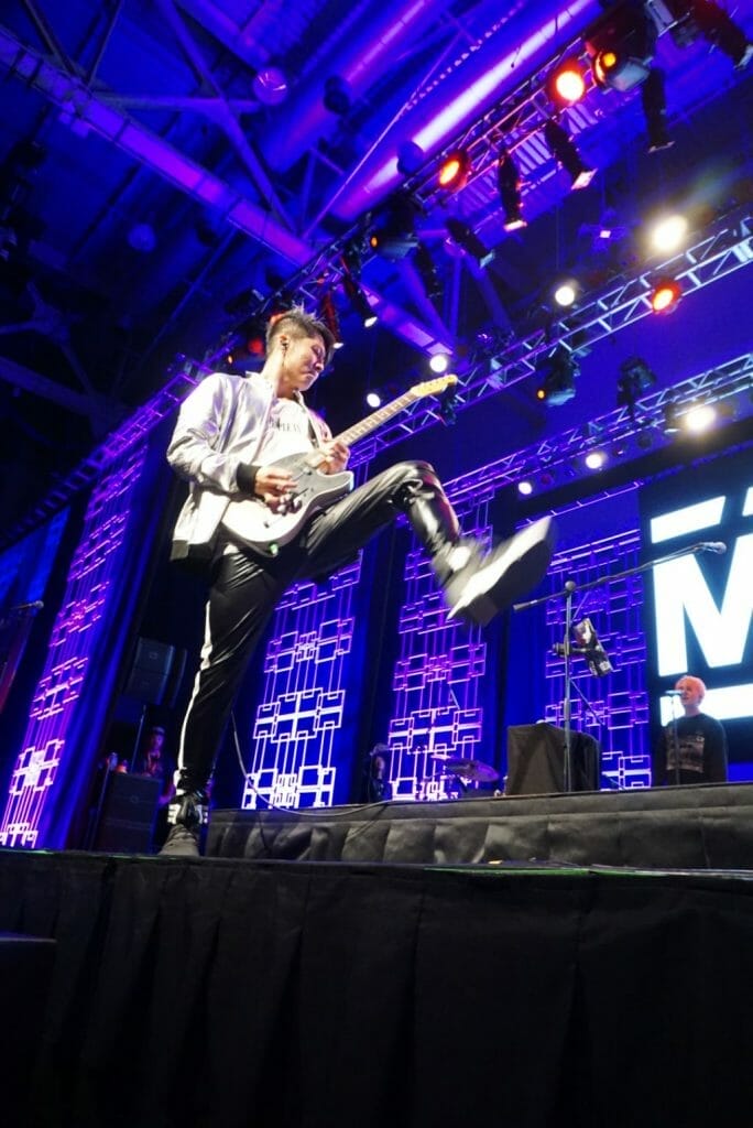 MIYAVI onstage at Anime Boston 2019, his foot coming down in a stomp. He's wearing a loose, white shirt and black pants with a white stripe along the side.