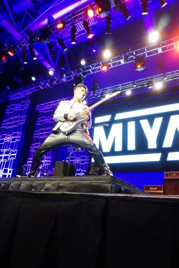 MIYAVI onstage at Anime Boston 2019, legs spread wide as he poses and plays his guitar. He's wearing a loose, white shirt and black pants with a white stripe along the side.