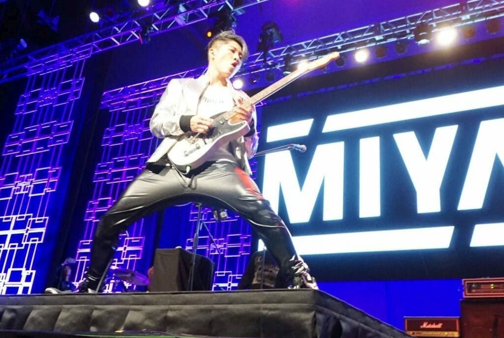 MIYAVI onstage at Anime Boston 2019, legs spread wide as he poses and plays his guitar. He's wearing a loose, white shirt and black pants with a white stripe along the side.