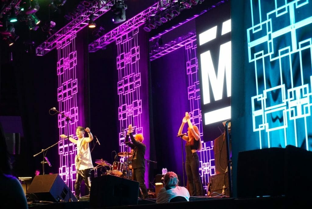 MIYAVI onstage at Anime Boston 2019, arms outstretched as backup singers clap along. He's wearing a loose, white shirt and black pants with a white stripe along the side.