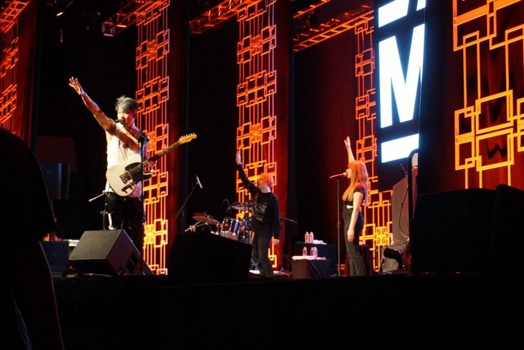 MIYAVI onstage at Anime Boston 2019. His right arm is outstretched as he sings.. He's wearing a loose, white shirt and black pants with a white stripe along the side.