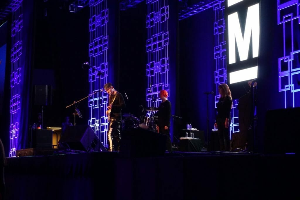Guitarist MIYAVI onstage at Anime Boston 2019, solemnly playing as backup vocalists stand behind him.. He's wearing a black shirt and black pants with a stripe running down the side.