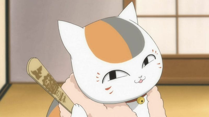 Natsume's Book of Friends Anime Still - a cat smirks playfully at the camera. It's sitting upright, wearing a towel on its neck and holding a Popsicle stick.