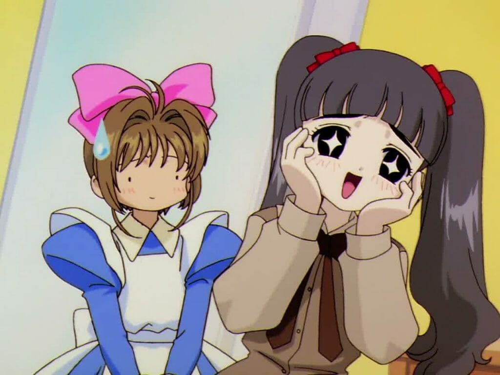 A black-haired girl with pigtails wears an elated expression as a brown-haired girl in a blue dress and pink bow stares, nonplussed.