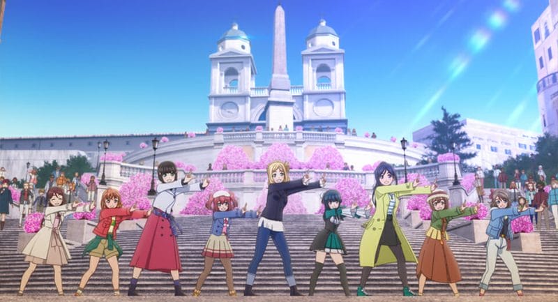Still from Love Live! Sunshine!! The School Idol Movie, which features nine women posing in front of a large white building.