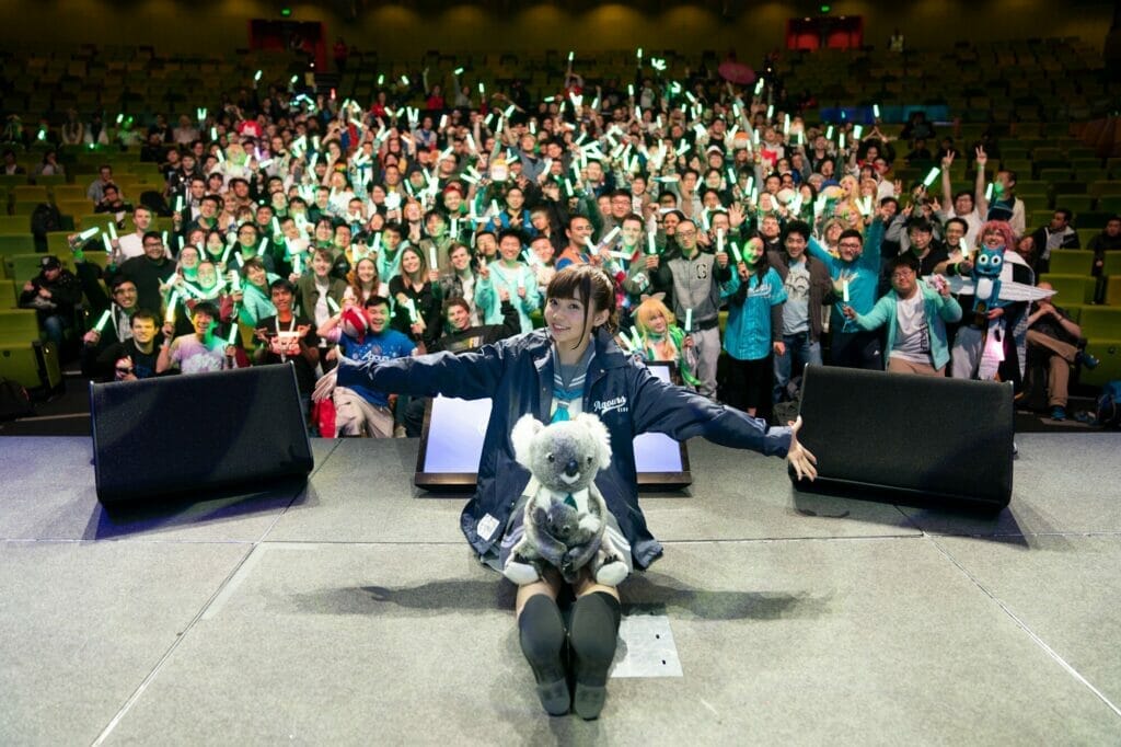 A Love Live voice actress sitting on stage with a koala plush in her lap. Behind her, an audience waves glowsticks