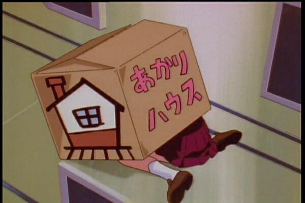 Still from Battle Athletes Victory (Original) - A young woman hides under a box with a crudely painted house and the words "Akari House" scrawled on it.