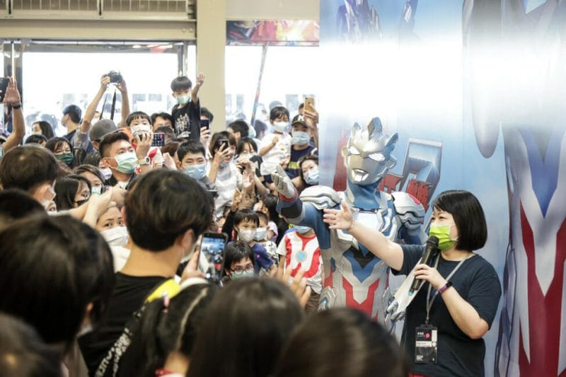 A number of people holding smartphones gather around a posing man in an Ultraman costume.