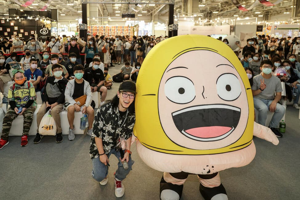 A man poses by a yellow mascot character, in front of an audience of onlookers.