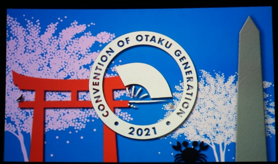 Photo of an illuminated sign that feature the Otakon logo set against a peaceful scene that has two cherry trees and a red shrine gate. A silhouette of the crab can be seen in the bottom-right.