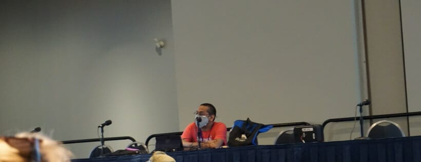 Daryl Surat, clad in an orange shirt and fac emask, sits at a table in front of a room filled with people at Otakon 2021.