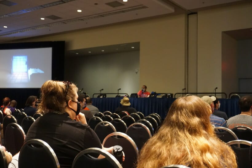 Daryl Surat, clad in an orange shirt and fac emask, sits at a table in front of a room filled with people at Otakon 2021.