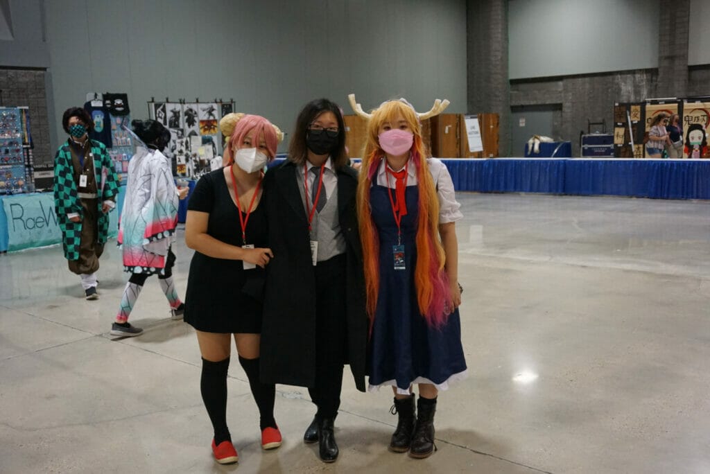 Photograph from Otakon 2021 - A dark-haired woman in a suit, a pink-haired woman in a black dress, and a blonde woman in a maid outfit pose for a photo.