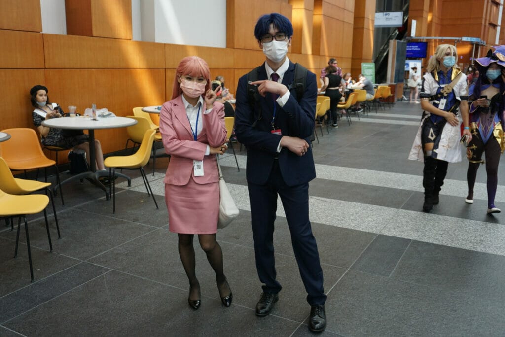 Photo from Otakon 2021 - A man in a blue suit and a woman in a pink business suit pose and point at each other playfully.
