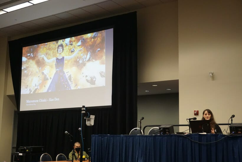 A brown-haired person sits behind a table with a blue tablecloth. A slide featuring Chiaki Mayumura is on a screen beside them.