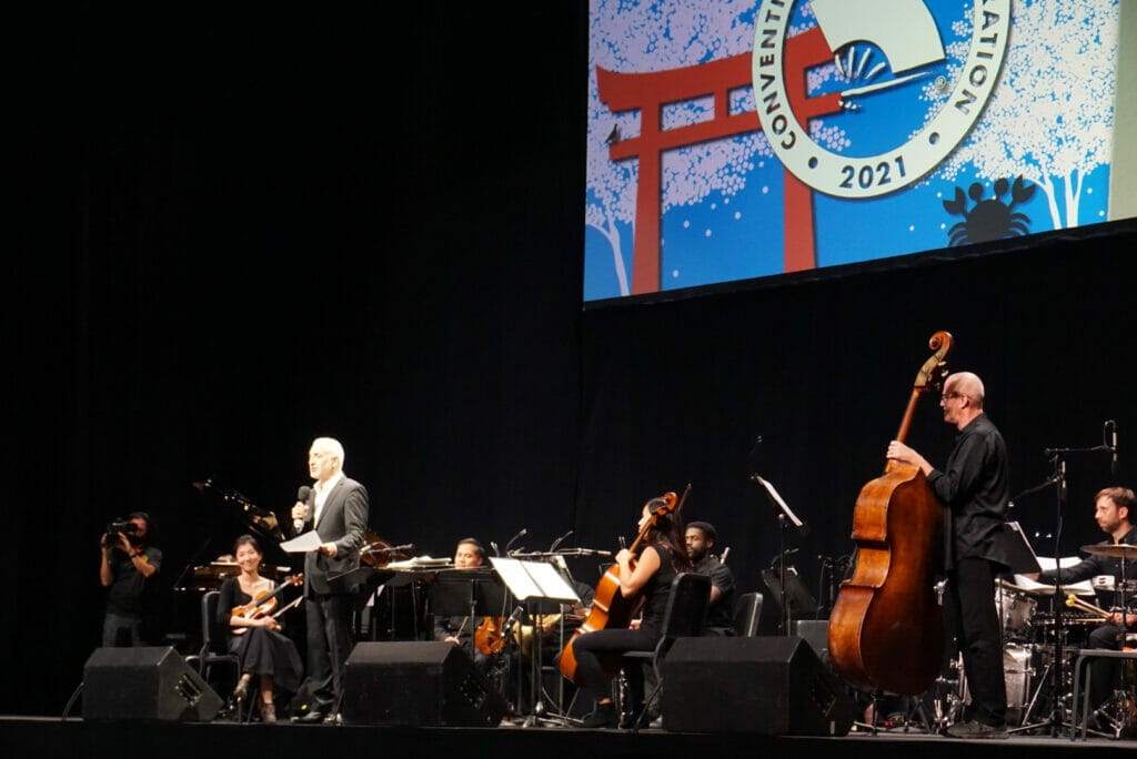 A silver-haired man in a suit stands onstage with a microphone, surrounded by musicians.