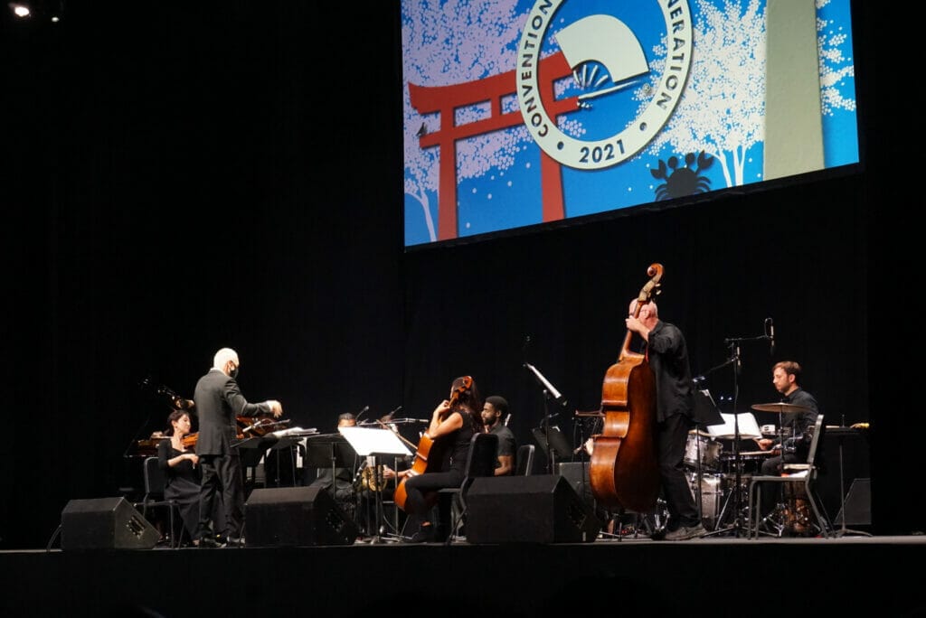 A silver-haired man conducts a small band of musicians beneath a large "Otakon 2021" sign.