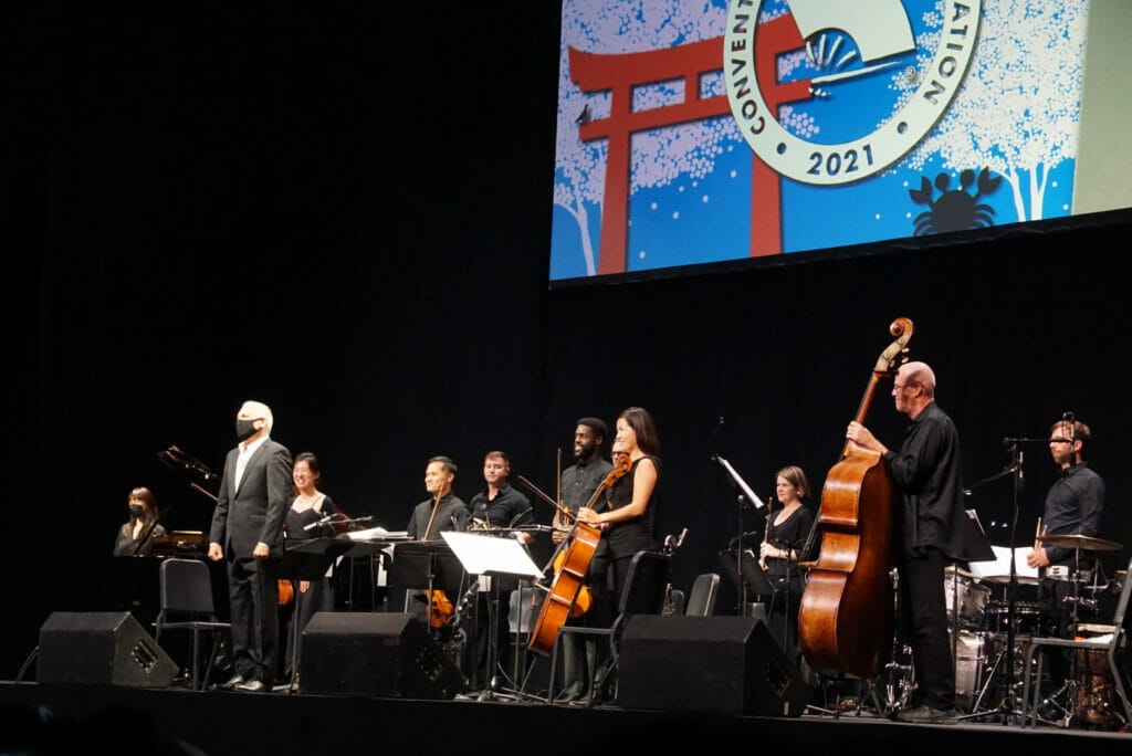 A silver-haired man in a black suit and face mask stands onstage with a microphone, surrounded by musicians. 