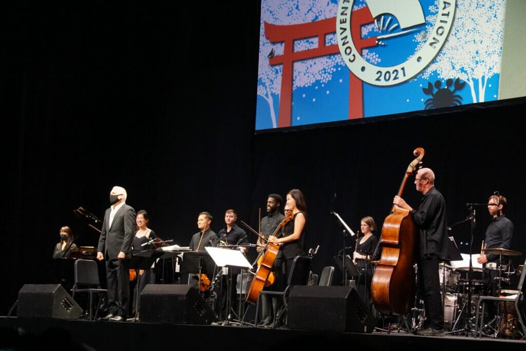 A silver-haired man in a black suit and face mask stands onstage with a microphone, surrounded by musicians.