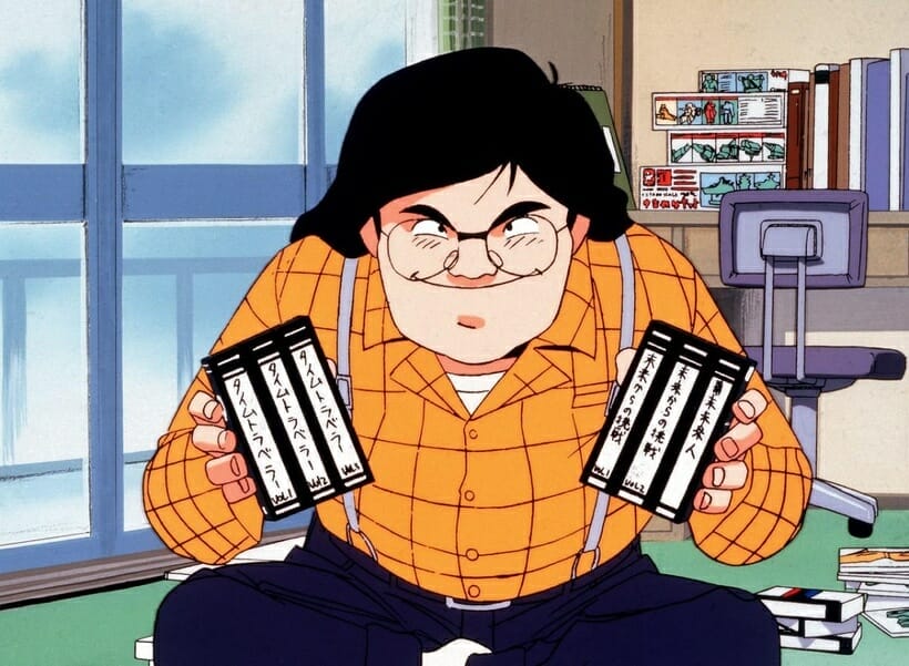 Still from Otaku no Video. A black-haired man in glasses and an orange shirt grins as he holds two fistfuls of VHS tapes.