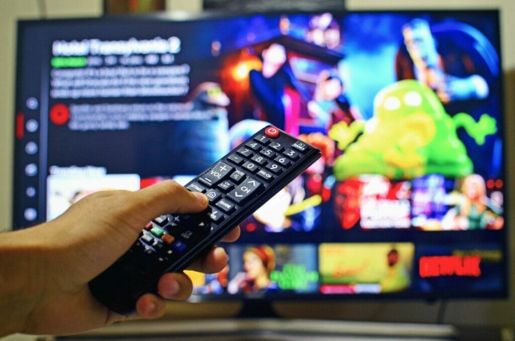 Close-up of a hand holding a TV remote. Behind it, a blurred image of a TV with Netflix up onscreen is visible.