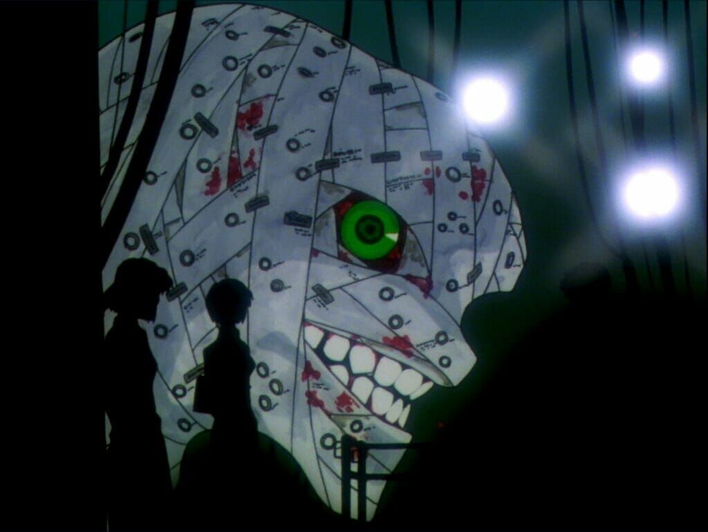 Side view of a giant creature, covered in bloody bandages, grinning as a light shines down upon it. Its green eye stares at the silhouettes of several humans in the foreground.