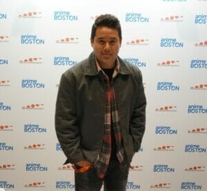 Actor Johnny Yong Bosch stands in front of a wall with the Anime Boston logo printed on it. He's wearing a brown coat