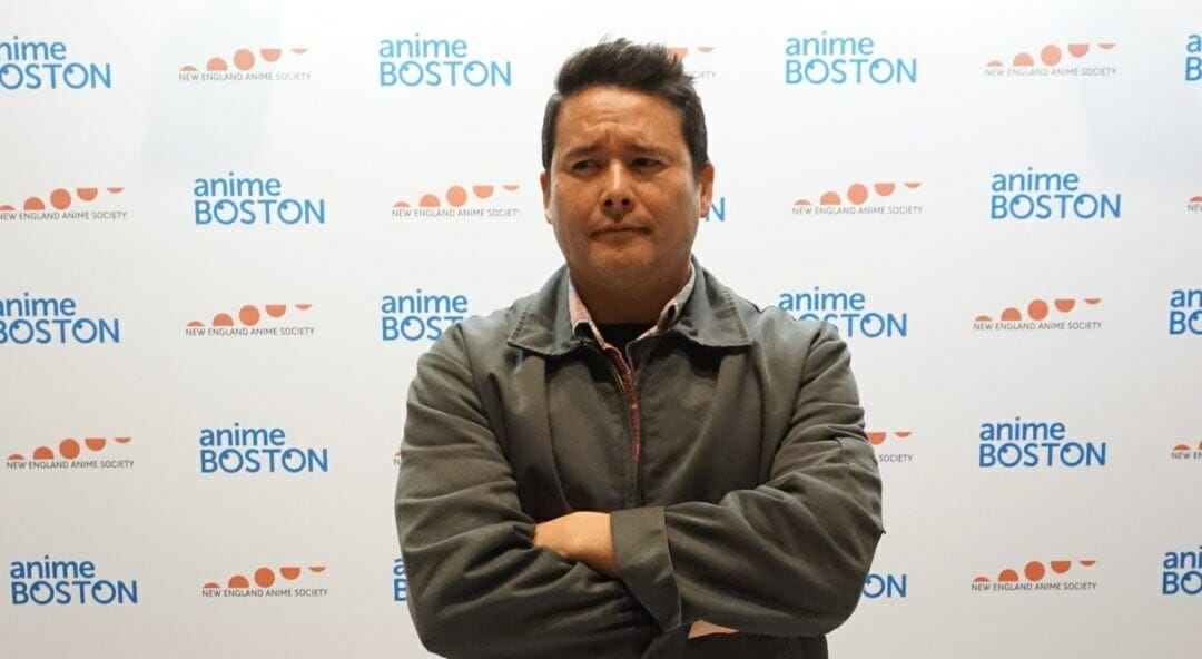 Headshot of Johnny Yong Bosch as he stands, arms folder, with a quizzical expression.