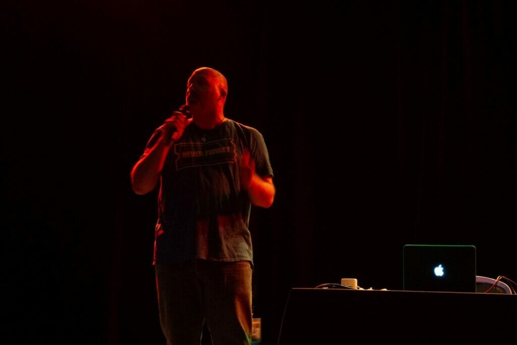 Photo from Anime Boston 2022: Brian T. Price addresses the crowd while bathed in a dim red light. His blue shirt features a picture of a ticket that reads "Never Forget"