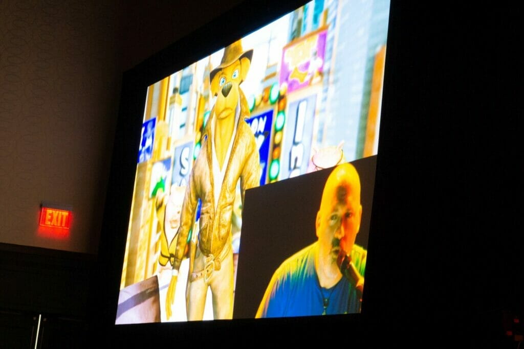 Photo of a screen with a cartoon dog projected on it. A video feed of a bald man talking into a microphone is in the bottom-right corner of the image,